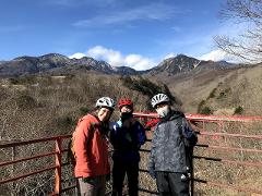 Embrace Beautiful Mother Nature on an E-Bike Tour of Yatsugatake Three people in cycling gear and helmets pose for a photo with a mountain landscape and a red bridge in the background.