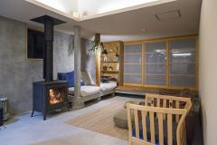 An interior shot shows a room with a modern rustic design, featuring a wood-burning stove, a concrete support, a neutral-toned sofa, and a natural fiber rug.