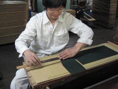 A man in a white shirt works with dark green fabric and a ruler-like tool on a wooden surface, with stacks of similar materials visible in the background.