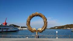 A golden laurel wreath sculpture stands on a waterfront promenade, framing a picturesque view of the sea, distant islands, and a passenger ferry under a clear blue sky.