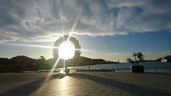 The sun shines through a circular, wreath-like sculpture on a paved waterfront, creating a striking silhouette and dazzling light.