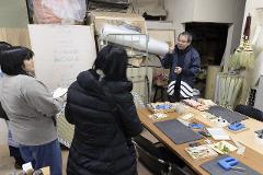 A group of people are in a workshop, looking at tatami mats and materials, with a craftsman explaining the process.