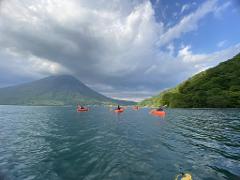 A group kayaks on a calm lake with a lush green mountain and a large volcano in the background under a partly cloudy sky.