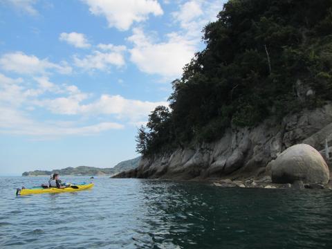 A person kayaking in a yellow kayak along a rocky coastline with trees, under a blue sky with scattered clouds.