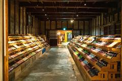 The image shows a sake tasting room with numerous bottles of sake displayed on tiered wooden shelves in a dimly lit, traditional Japanese-style building.