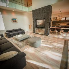 A modern living room features a gray sofa, textured rug, a stone-clad media center, and open layout to a dining area with a chandelier.