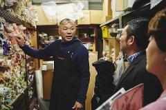 A man in an apron points to a wall of colourful fabrics while two men in suits look on in a workshop.