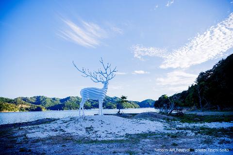 A white, stylized deer sculpture stands on a hill overlooking a body of water and tree-covered mountains under a bright blue sky.