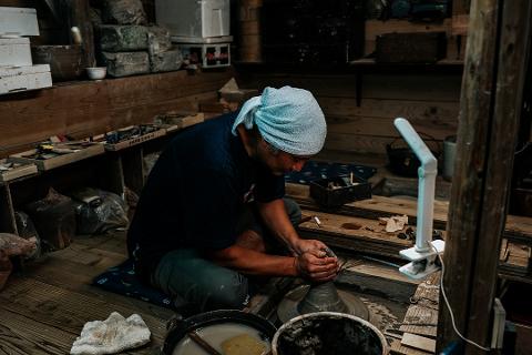 Pottery & Porcelain: Stories of the Artisans Behind the Craft A potter works at a wheel in a rustic studio, his hands shaping clay as he is illuminated by a modern desk lamp.