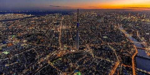 An aerial view of Tokyo at dusk, with the Tokyo Skytree illuminated and city lights twinkling below as the sun sets over the horizon.