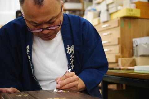 An artisan in a blue garment and glasses works on a wooden surface with a tool, surrounded by craft supplies and wooden cabinets.