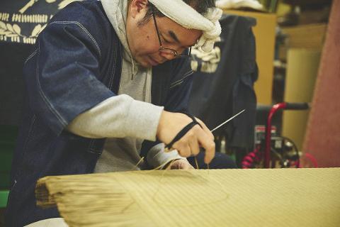 A craftsman with a towel on his head works on a tatami mat, using a needle to weave straw.