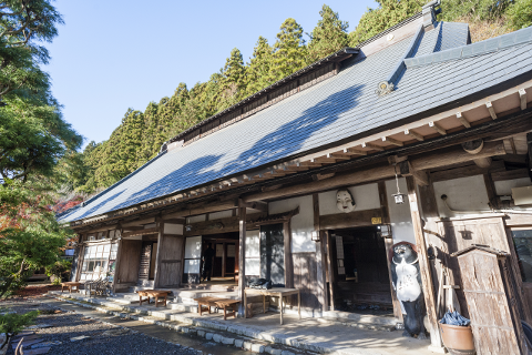 A traditional Japanese building with a tiled roof and wooden balconies stands against a backdrop of lush green trees under a clear blue sky.