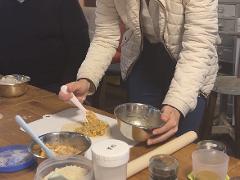 A person in a quilted jacket fills a pastry with filling from a stainless steel bowl using a pink spoon, surrounded by ingredients and cooking utensils on a wooden table.
