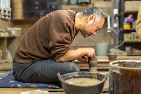 Ceramică și porțelan: Povești ale artizanilor din spatele meșteșugului A group of people look on as a man sits and works on pottery in a rustic studio.