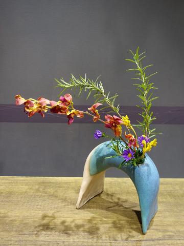 A flower arrangement in an arch-shaped vase features colorful orchids, yellow blossoms, purple asters, and rosemary sprigs.