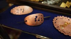 A copper hair accessory with cherry blossom cutouts and another piece of copper metalwork with a hanging jewel sit on a blue surface.