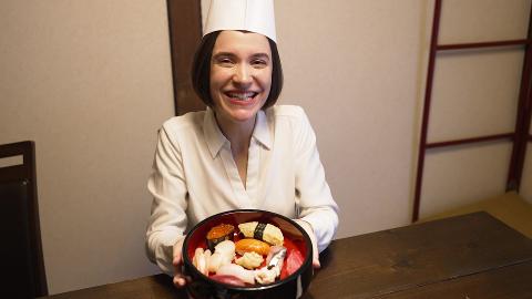 A smiling woman wearing a chef's hat and white shirt sits at a wooden table, holding a bowl of sushi and other ingredients.
