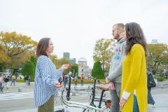 A tour guide is talking to a couple with bicycles at the Hiroshima Peace Memorial Park, with the Atomic Bomb Dome visible in the background.