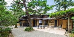 A tranquil Japanese garden setting surrounds a modern building with outdoor seating, featuring large windows and a traditional wooden facade.