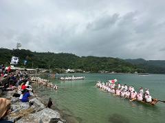 A crowd watches longboats race on Shioya Bay in Okinawa, Japan, under a cloudy sky, with a Japanese flag waving from one of the boats.
