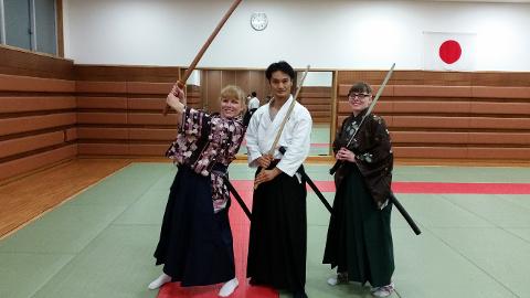 Traja ľudia, dve ženy a jeden muž, v tradičnom japonskom oblečení pózujú s mečmi v dojo bojových umení s japonskou vlajkou v pozadí.