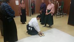 Skupina ľudí oblečených v tradičnom japonskom oblečení sa zhromaždila v interiéri, pričom jeden z nich kľakol a položil si sandále na bielu podložku.