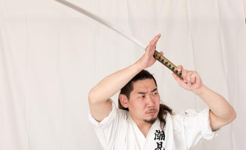 A man in a white karate uniform wields a katana sword overhead, looking intently to the side with a focused expression against a plain backdrop.