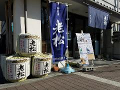 A storefront features sake barrels, blue banners with Japanese script, and a sign displaying prices and information.