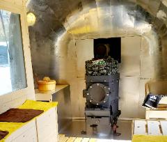 This is an interior shot of a sauna, featuring a black stove filled with rocks, wooden benches with yellow towels, and a bucket with a wooden ladle. The interior is lined with reflective metal panels.