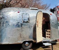 A shiny Airstream trailer with an open wooden door revealing a sauna setup inside, parked outdoors on a sunny day.