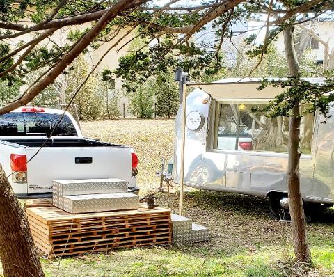 A white pickup truck is parked next to a silver Airstream sauna trailer in a grassy area with trees.