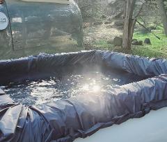 The image shows a makeshift outdoor hot tub made with a tarp liner, reflecting sunlight, next to a vintage Airstream trailer in a grassy yard.