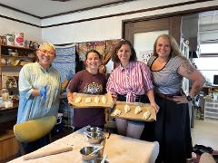 Uma pessoa com uma jaqueta acolchoada recheia um pastel com recheio de uma tigela de aço inoxidável usando uma colher rosa, cercada por ingredientes e utensílios de cozinha em uma mesa de madeira.