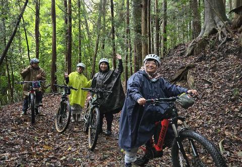 A large group of people with bicycles in a grassy area with trees and mountains in the background.