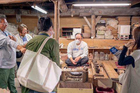 Pottery & Porcelain: Stories of the Artisans Behind the Craft A group of people look on as a man sits and works on pottery in a rustic studio.