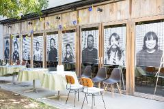 Banners and portraits of people are displayed outside a building with wooden-paneled walls and glass doors, with tables and chairs set up in front.