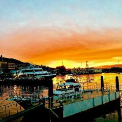 A picturesque harbor scene at sunset, featuring a large yacht and a small boat docked near a pier, with a vibrant orange and blue sky reflecting on the water.