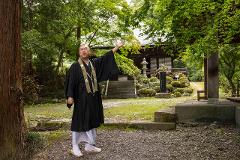 A Buddhist monk gestures while standing in front of a temple in a serene, wooded setting.