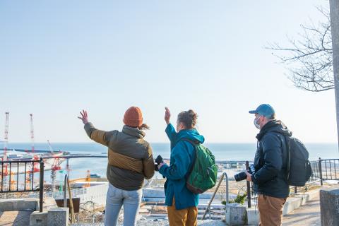 Three people stand on a hilltop overlooking a harbor with construction cranes and the ocean, suggesting a tour focused on disaster recovery and heritage.
