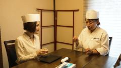 Two women, wearing chef hats and white shirts, sit at a wooden table, one gesturing while the other listens, in a brightly lit room, ready to make sushi.