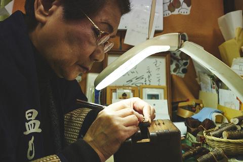 An artisan carefully carves a traditional Japanese hanko seal using natural wood under a bright lamp.