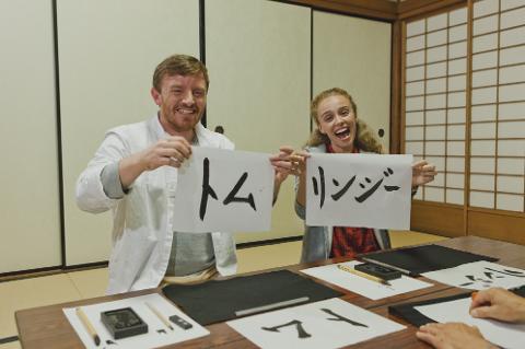 Two people gleefully hold up papers with Japanese calligraphy of their names while seated at a table with calligraphy supplies.