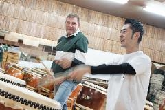 Two men play taiko drums in a studio with many drums and wooden boxes stacked in the background.
