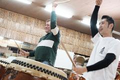 Two men practice playing the Taiko drums in front of a wall adorned with wooden plaques bearing Japanese calligraphy.