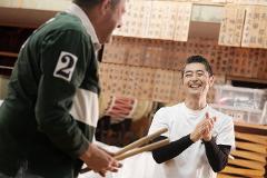 Two men are in a room with Japanese calligraphy on the walls, one is wearing a green rugby shirt with a number 2 on it and holding drumsticks, while the other is wearing a white t-shirt and clapping his hands.