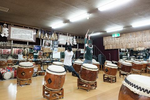Nagano Morning Taiko Drumming – Private Dojo & Museum Tour Two men with drumsticks practice playing large traditional Japanese drums in a studio filled with more drums and artwork.