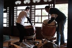 Two men in a traditional Japanese room are preparing to play large taiko drums.