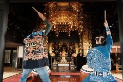 Two men in traditional Japanese attire play taiko drums in a temple, with a golden altar and ornate decorations in the background.