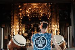 A taiko drummer stands ready to play in a brightly lit, ornately decorated Japanese temple.